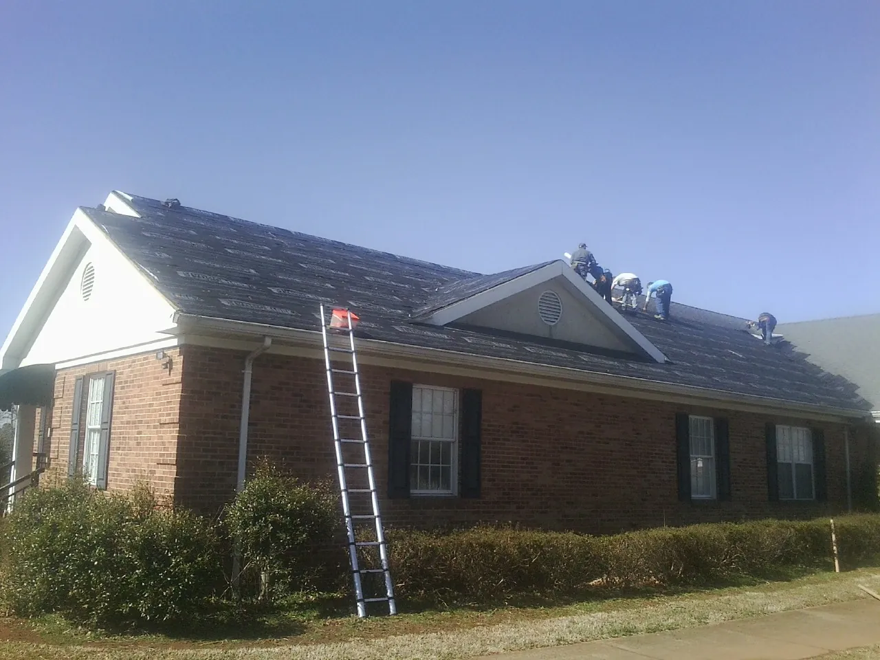 Crew tearing shingles off a brick ranch roof with a ladder set — Metal Roof Repair work by our crew in North Chili
