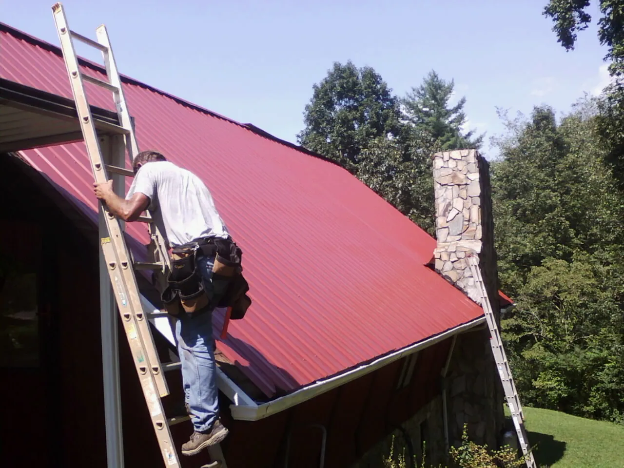 Roofer on a ladder beside a red metal roof and stone chimney — Soffit & Fascia Repair by Roofing Brothers in North Chili